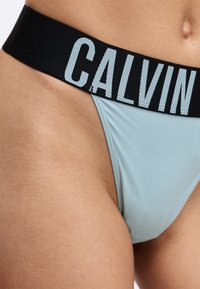 Lichtblauwe string met een zwarte elastische tailleband, met 'CALVIN' in grote, lichtblauwe letters. Glad textuur, ontwerp met minimale bedekking.