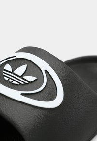 Crna natikača s teksturiranim remenom ukrašenim bijelim logotipom i okruglim detaljem, ravnim potplatom i minimalističkim dizajnom.