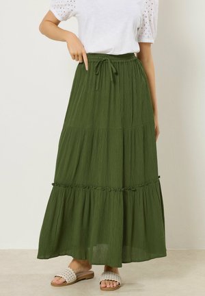 Frau trägt eine weiße Bluse und einen olivgrünen Maxirock mit Kordelzugbund, steht barfuß auf einem hellen Boden.