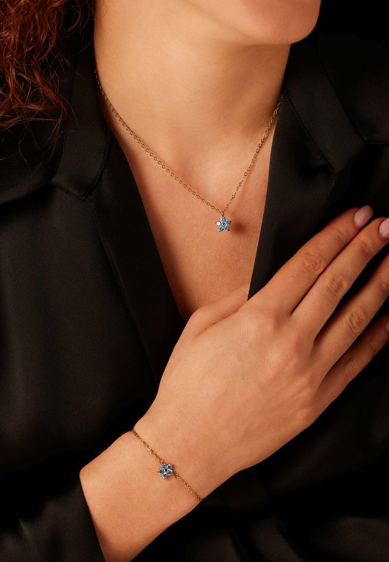 Gouden halsketting en armband set met blauwe bloempendants met ingewikkelde details. De halsketting hangt vlakbij het sleutelbeen, de armband om de pols.
