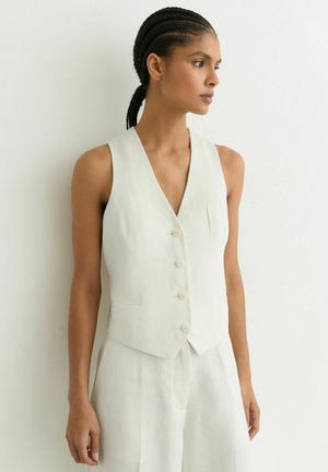 Žena s vrkočmi, oblečená v bielu zapínateľnú vestu a zodpovedajúce nohavice, stojí a pozerá sa doprava na jednoliatom pozadí.