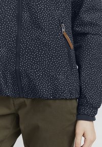 Giacca navy con pois bianchi, chiusura con zip e polsini a coste. Presenta una tasca laterale con un accento in pelle. Tessuto liscio e leggero.