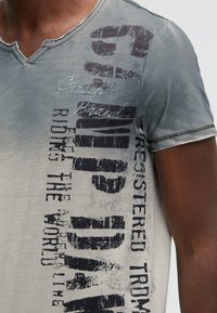 Gray cotton T-shirt with a V-neck, featuring distressed edges and bold printed text in black and dark gray tones on the front.