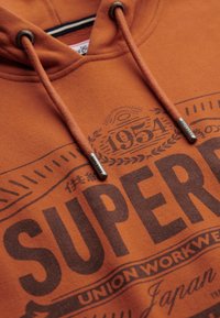 Pomarańczowy hoodie z kapturem na sznurki, z wyraźnym nadrukiem "SUPERDRY", teksturowanym materiałem i sznurkami z metalowymi końcówkami.