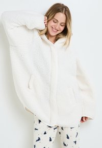 Giacca oversized in pile bianco con chiusura frontale, ampie tasche e una consistenza morbida. Abbinata a pantaloni da pigiama a fantasia con gatti.