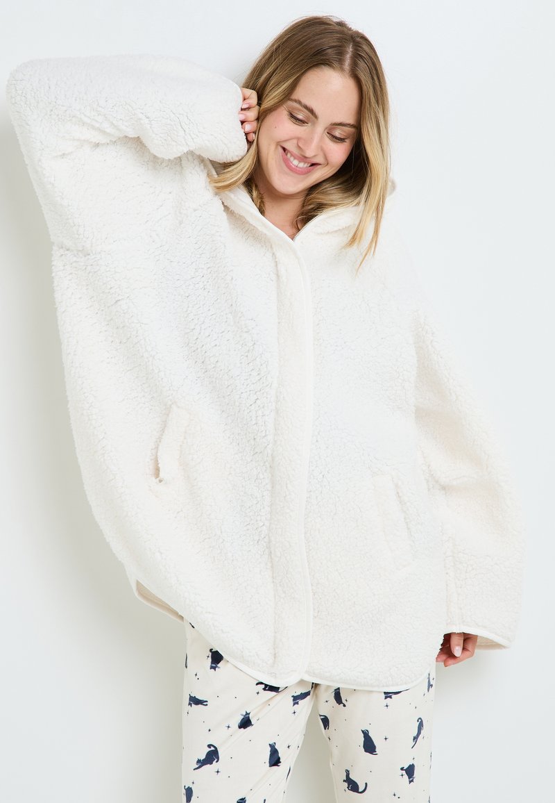 Giacca oversized in pile bianco con chiusura frontale, ampie tasche e una consistenza morbida. Abbinata a pantaloni da pigiama a fantasia con gatti.