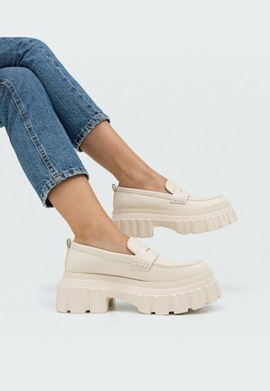 Fødder iført off-white chunky plateau loafers kombineret med op rullede blå jeans mod en ensartet lys baggrund.
