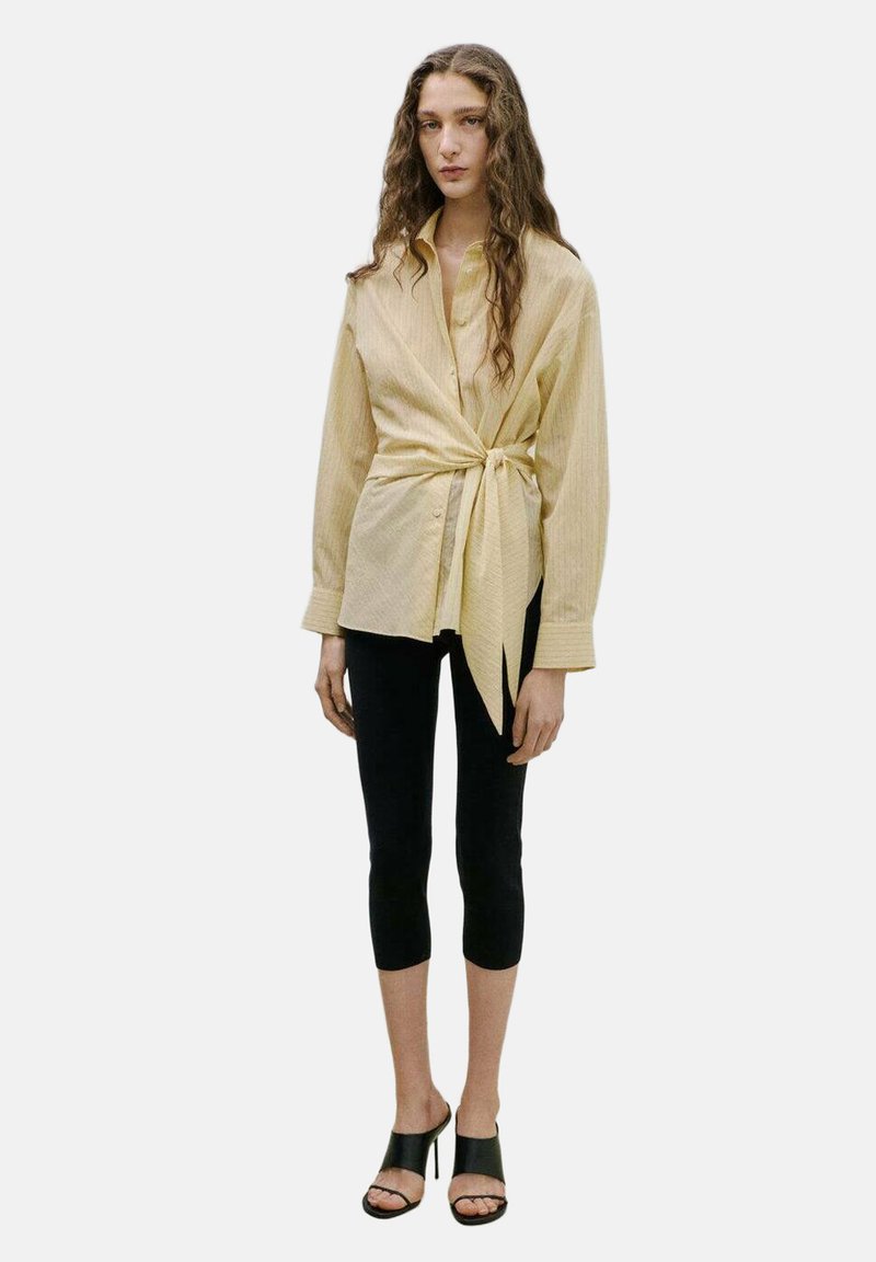 Ženska z dolgimi valovitimi lasmi, oblečena v bež srajco z gumbi in ovitkom spredaj, črne kratke legice in črne sandale z odprtimi prsti in visokimi petami.