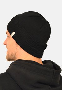 Black knitted beanie hat with a snug fit, featuring a ribbed texture and a small white label on the side. Worn with a black hoodie.