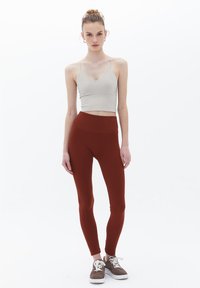 Světle šedý crop top s tenkými ramínky, doplněný oleggings vysokého pasu v barvě mahagonu. Modelka stojí s rukama podél těla na bílém pozadí.