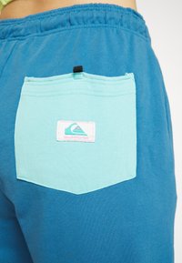 Blue sweatshorts with a large, light blue patch pocket featuring a pink Quiksilver logo. Elastic waistband and soft texture.