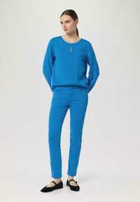 Jasnoniebieska bluzka z dekoltem w kształcie łezki i długimi rękawami, połączona z dopasowanymi, niebieskimi spodniami typu slim-fit oraz czarnymi płaskimi butami, na neutralnym tle.