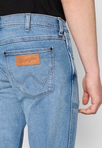 Heledad sinine denim, millel on nahast Wrangler'i silt, viie tasku disain ja kontrastsete õmbluste detailid.