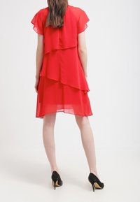 Robe en chiffon rouge avec un design superposé, présentant des manches courtes et une silhouette fluide. Portée avec des chaussures à talons hauts noires.