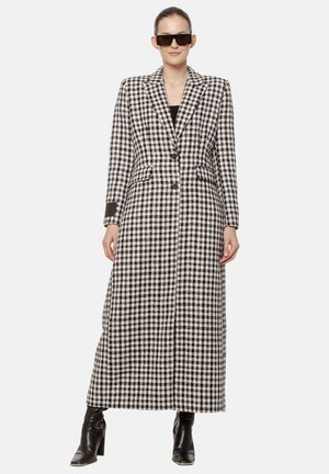 Kobieta w długim czarno-białym w kratę płaszczu, czarnych botkach i dużych czarnych okularach przeciwsłonecznych, stojąca na jednolitym tle.