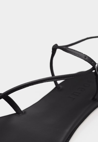 Μαύρο επίπεδο σανδάλι με λεπτές δερμάτινες λουριά και μινιμαλιστικό σχέδιο σε λευκό φόντο.