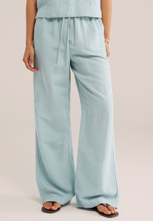 Vrouw in lichtblauwe wijde broek met trekkoord in de taille en zwarte teenslippers, staand tegen een effen achtergrond.