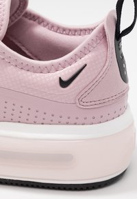 Ljust rosa träningsskor med ett texturerat tygöverdrag, vit mellansula, svart Nike-logga och cirkulära perforeringar på sidan. Svart hälflik.