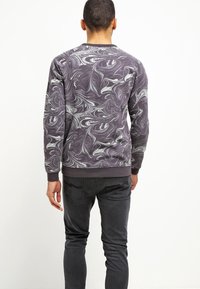 Man met een donkergrijs en wit gemarmerd shirt met lange mouwen en zwarte jeans, staand met de rug naar de camera tegen een witte achtergrond.