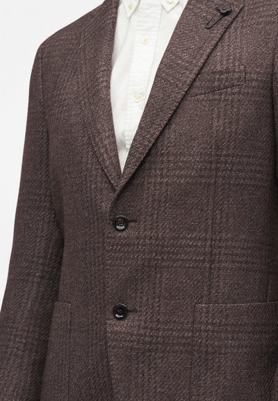 Blazer marron texturé avec un motif tartan subtil, boutons noirs et une épingle de revers noire en forme de fleur, porté sur une chemise blanche à boutons.