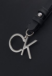 Yksityiskohta mustasta, teksturoidusta nahkabagista, jossa on hopeanvärinen CK-logo-riipus mustassa hihnassa, jossa on metallinen napituskoriste.