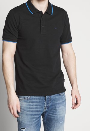 Homme portant un polo noir avec une bordure bleue et un jean bleu clair usé, debout devant un fond blanc uni.