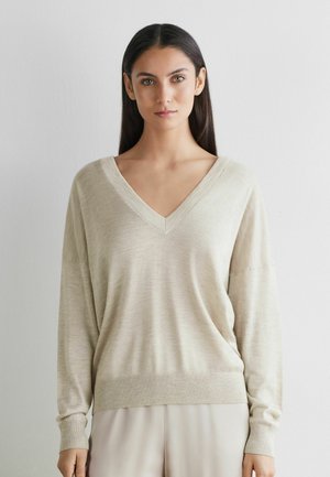 Jeune femme aux longs cheveux bruns portant un pull beige à col en V et un pantalon clair, debout contre un mur blanc uni.