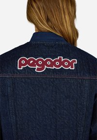 Τζάκετ τζιν σε σκούρο μπλε με υφή. Διαθέτει μεγάλο κόκκινο κεντημένο λογότυπο "pegador" στην πλάτη, με λευκό περίγραμμα από ραφές.