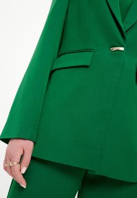 Blazer verde su misura con un colletto affilato, due tasche laterali e un elegante bottone in accento dorato. Realizzato in tessuto testurizzato.