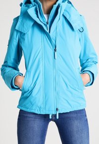 Blauw waterdicht jack met een dubbele capuchon, ritssluiting aan de voorkant en twee zijzakken. Heeft een gladde textuur en verstelbare koordjes aan de zoom.