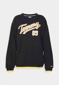 Svart långärmad sweatshirt med gul "Tommy 85" text på framsidan, gul kantning på ärmslut och nederkant, samt liten logotyp på vänster ärmslut.