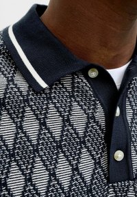 Dark-skinned person wearing a navy blue and white patterned knit polo shirt with a white undershirt visible at the collar.