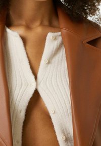 Slonovinový pletený svetr s žebrovanou strukturou a malými knoflíky, vrstvený pod hnědou koženou bundu se strukturovaným límcem.