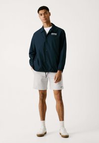 Chaqueta cortavientos azul marino con botones a presión, cuello y puños elásticos, combinada con pantalones cortos grises y zapatillas blancas, de pie sobre un fondo neutro.