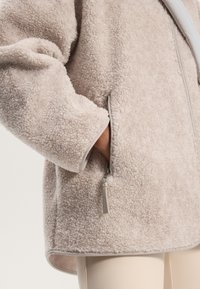 Lys beige fleecejakke med en tekstureret overflade, der har en lynlåslomme og kontrasterende grå kant langs kanten af ærmerne.
