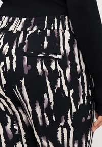 Žena v černobílých pruhovaných kalhotách s elastickým pasem a kapsou na zadní straně, v kombinaci s černým topem s dlouhým rukávem.