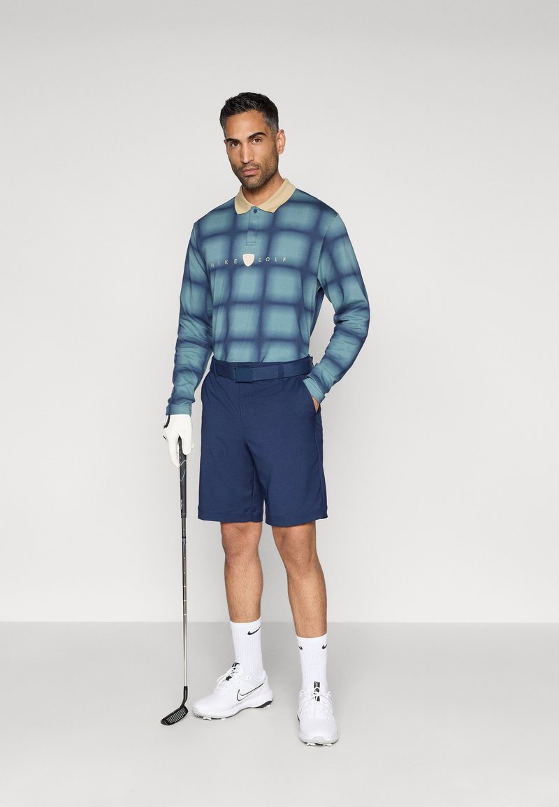 Langärmeliges türkisfarbenes Poloshirt mit Rastermuster, beigem Kragen, Logo auf der Brust, kombiniert mit marineblauen Shorts, weißen Sneakers und einem Golfschläger.