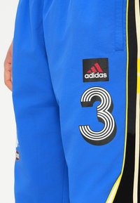 Zili sporta šorti zilā krāsā ar izteiktu melnbalto svītraino skaitli "3" grafiku, adidas logo uzšuvumu, dzeltenām un melnām sānu akcentiem.