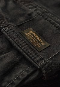 Czarna tkanina dżinsowa zbliżenie na zieloną metkę z napisem "SUPERDRY", z złotym tekstem i przeszyciami wzdłuż szwu kieszeni.