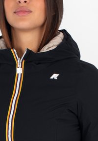 Črna jakna z sprednjim zadrgo, večbarvnimi črtami, logotipom na prsih in kontrastno bež podlogo v kapuci.