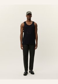 Svart strikket tanktopp med et teksturert design, kombinert med mørkegrå bukser og svarte sko. Modellen har på seg en grønn cap.