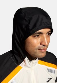 Veste à capuche avec une capuche noire, un corps blanc et des accents orange. Elle possède une fermeture éclair et des détails réfléchissants sur une texture lisse.
