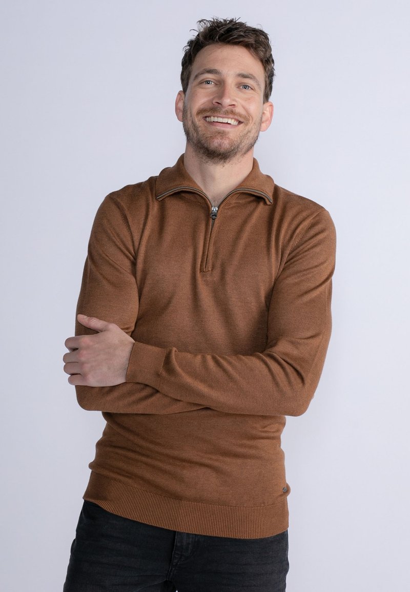 Brun strikket sweater med halv-zip krave og ribbede manchetter og kant. Bæret over mørke jeans, med en subtil tekstur og en tætsiddende pasform.