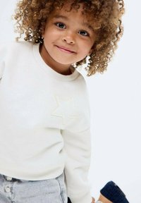 Valkoinen collegepaita, jossa on teksturoitu tähtikuvio rinnassa, yhdistettynä harmaisiin farkkushortseihin. Mallilla on kiharat hiukset ja neutraali ilme.