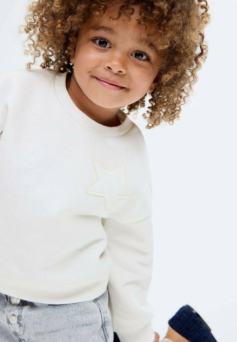 Valkoinen collegepaita, jossa on teksturoitu tähtikuvio rinnassa, yhdistettynä harmaisiin farkkushortseihin. Mallilla on kiharat hiukset ja neutraali ilme.