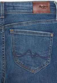 Vaqueros azules de mezclilla con un parche trasero de cuero con el logo, costuras en contraste y un patrón decorativo de ondas en el bolsillo trasero.