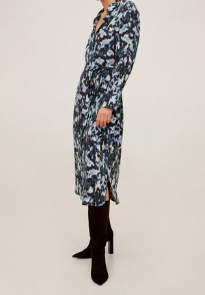 Kobieta w długiej sukience midi z długim rękawem, z paskiem, w niebiesko-czarny wzór, z bocznym rozcięciem, w połączeniu z czarnymi, sięgającymi kolan, botkami na obcasie.