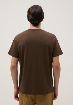 Uomo con capelli corti e scuri di spalle, indossa una maglietta marrone semplice a maniche corte e pantaloni color senape su sfondo neutro.