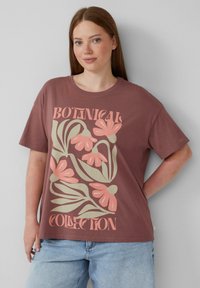 T-shirt en coton marron avec un design floral en rose et vert portant le texte "COLLECTION BOTANIQUE". Manches courtes et coupe décontractée.