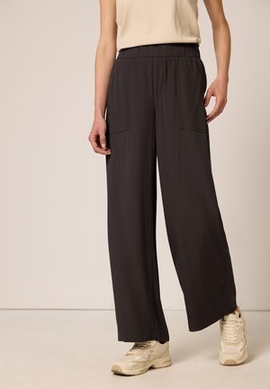 Frau trägt lockere schwarze Hose mit vorderen aufgesetzten Taschen, beige ärmelloses Oberteil und beige Sneaker, steht vor neutralem Hintergrund.